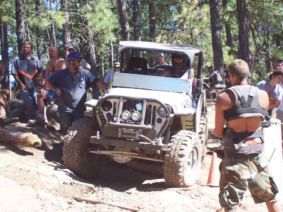 White CJ-style Jeep with diamond-plate fenders, soft top and winch bumper surrounded by crowd on dusty trail