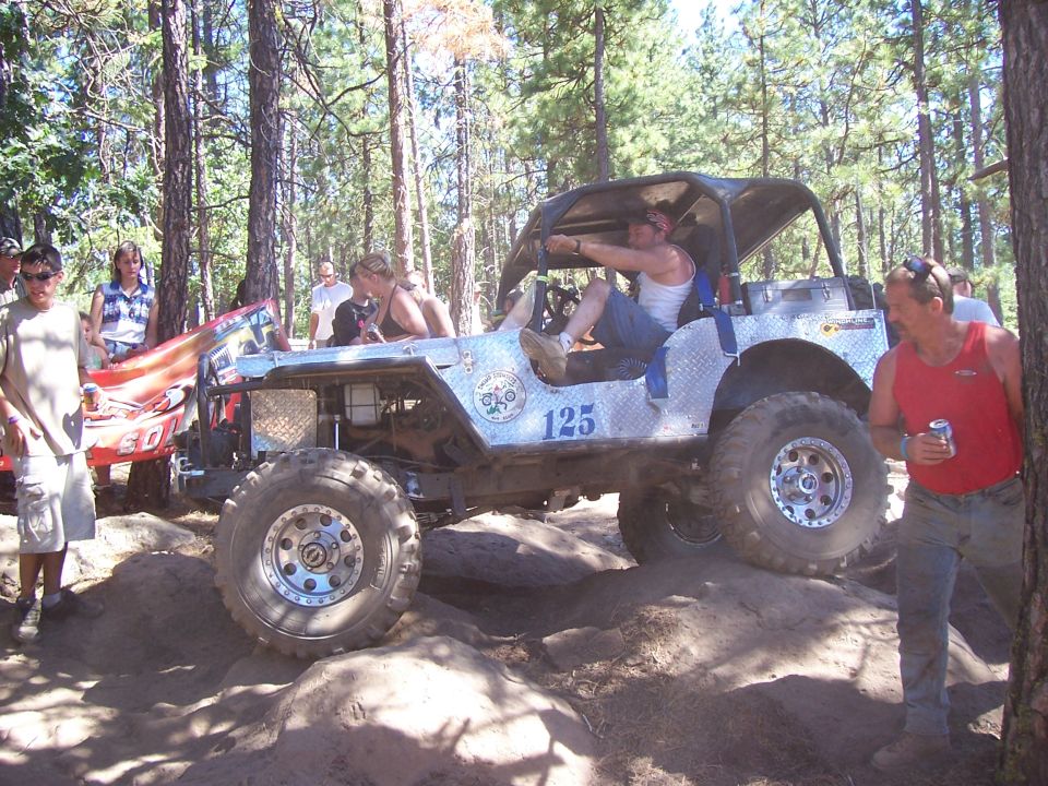 Diamond-plate Jeep number 125 with driver inside parked on dirt, spectators and man with beer can beside it