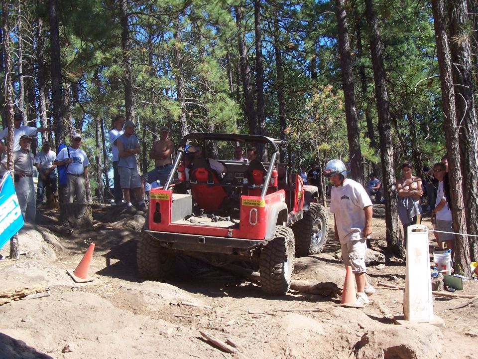 Red CJ Jeep on a dirt obstacle facing a crowd of spectators in a pine forest, cones marking the line
