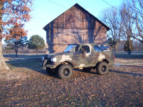 1988 Ford Bronco - A camouflaged Ford Bronco sits in a grassy field with a rustic barn in the background.
