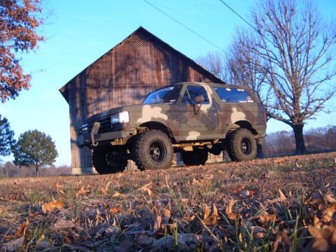 1988 Ford Bronco - A camouflaged Ford Bronco sits on a dry, grassy field with a rustic barn in the background under a clear blue sky.