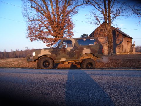 1988 Ford Bronco - A camouflaged Ford Bronco sits parked on the side of a road with a rustic barn in the background.
