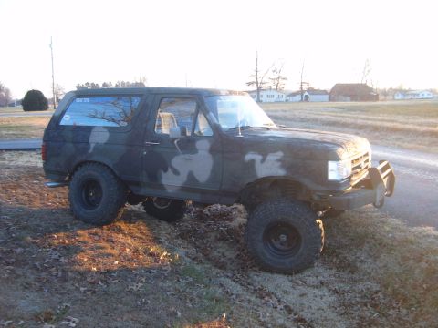 1988 Ford Bronco - This lifted Ford Bronco, painted in camouflage, sits on a dirt embankment under a bright sky.