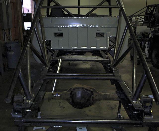 Truck chassis with custom roll cage and rear axle visible. Bed of truck is grey