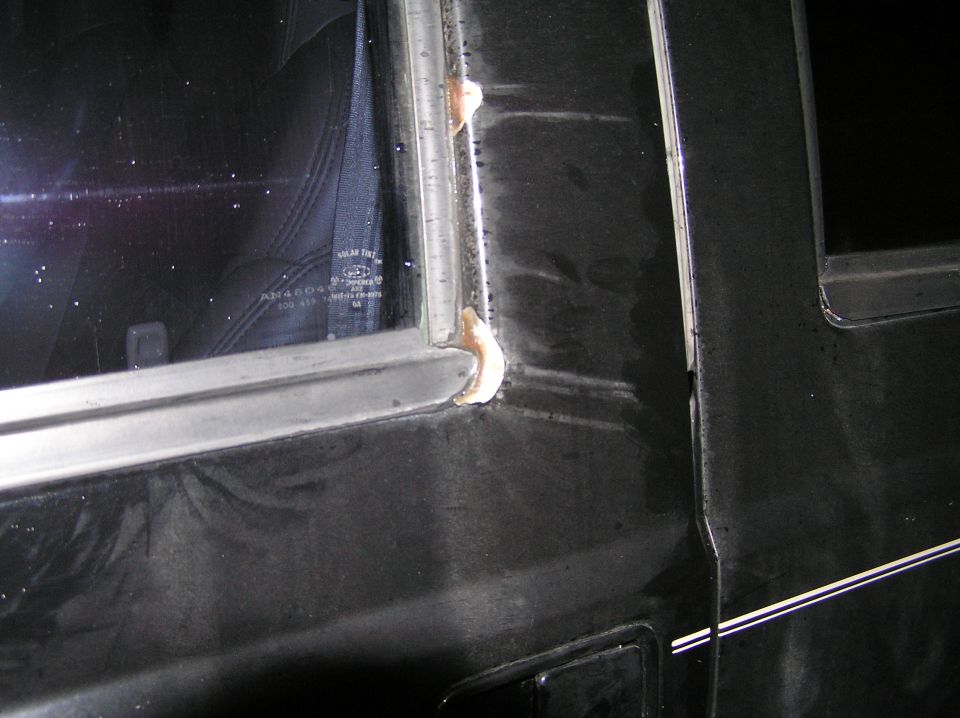 Black vehicle window frame corner with tan sealant globs and tinted glass etching