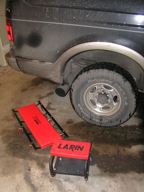2000 Ford Excursion - Rear view of a dark SUV with a red creeper and stool nearby, suggesting maintenance work.