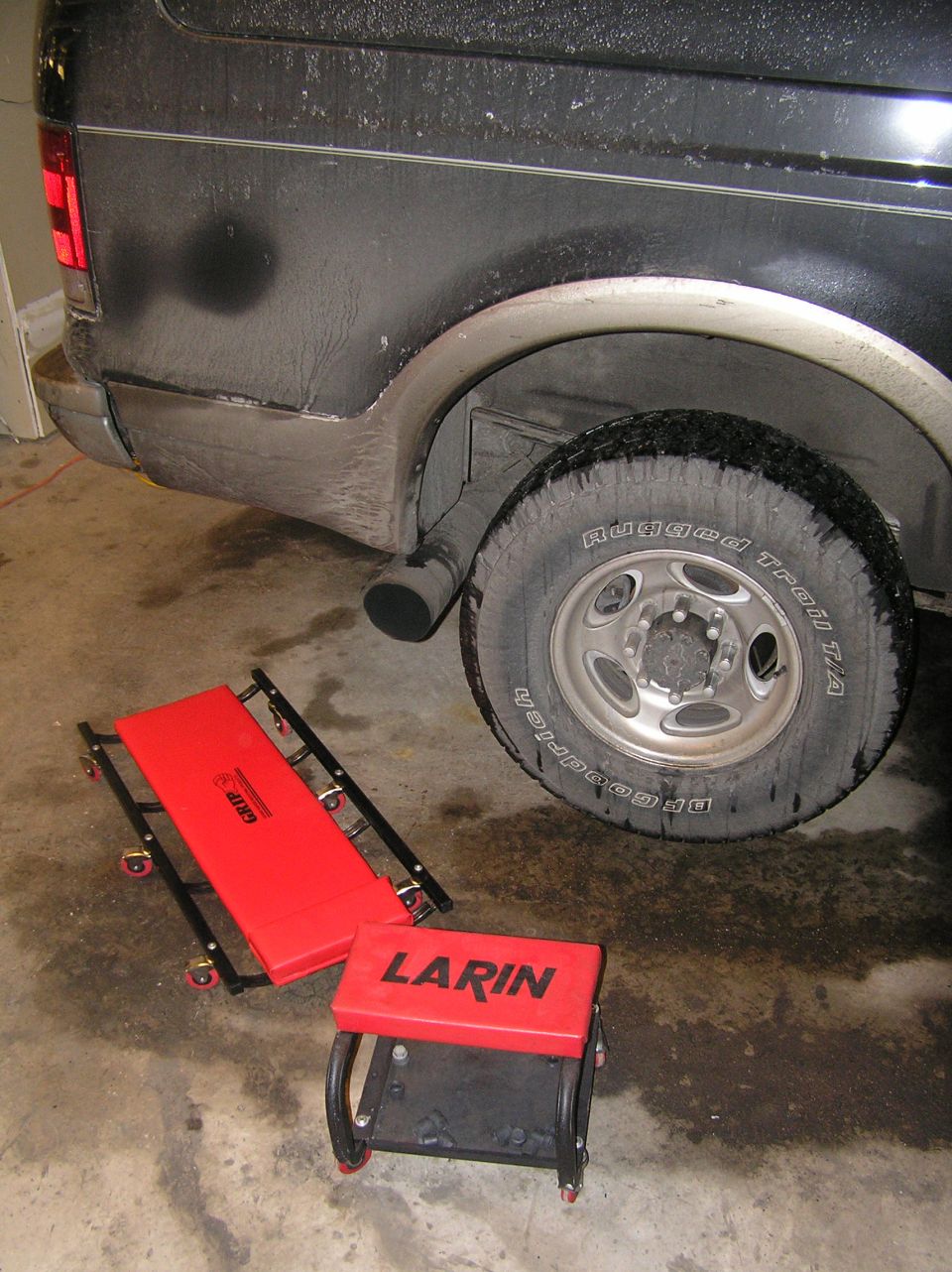 Black SUV rear wheel above red creeper and LARIN mechanic's stool on garage floor