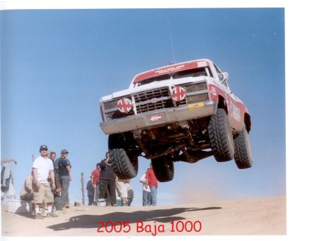 1990 Ford Bronco - white and red off-road race truck catches air over a desert berm as spectators watch; caption reads 2005 Baja 1000.