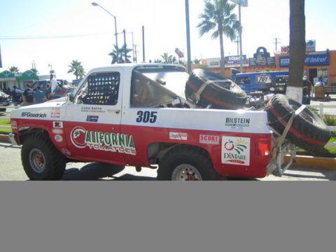 1990 Ford Bronco - Side rear view of a white and red off-road race truck number 305 carrying two spare tires, parked along a palm-lined street with shops behi…