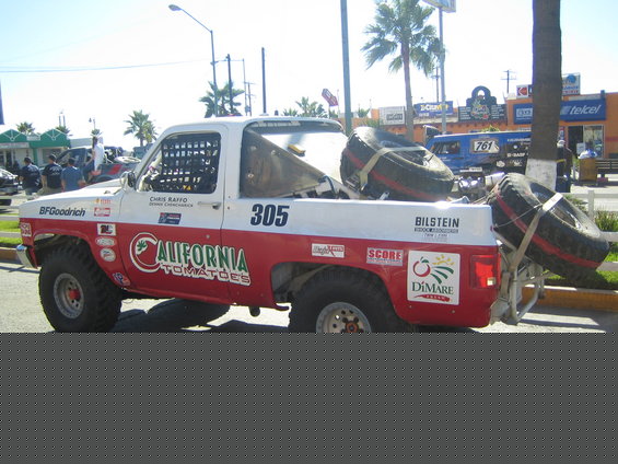 Side rear view of white and red race truck #305 with spare tires on a palm-lined street