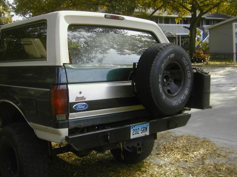 1993 Ford Bronco - Rear three-quarter view of a green Ford Bronco with a white hardtop, fitted with a heavy black steel rear bumper, swing-out carrier holding…