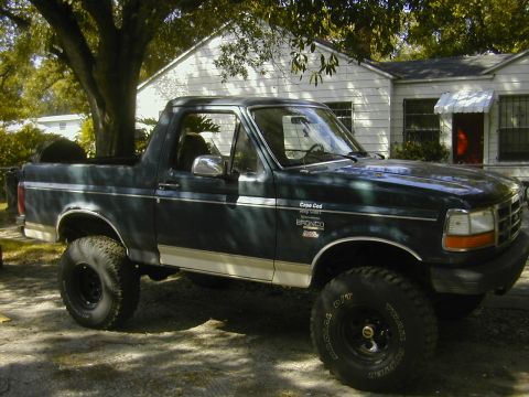 1993 Ford Bronco - Passenger-side profile of a lifted dark green Ford Bronco with its rear hardtop removed, parked in a shaded driveway beside a white house.…