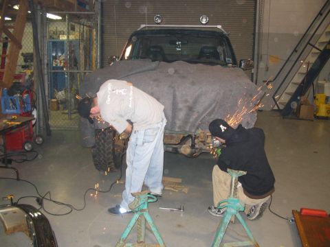 1996 Ford Bronco - Two individuals are actively working on the front of a vehicle, which is partially covered by a large gray tarp, with sparks flying from th…