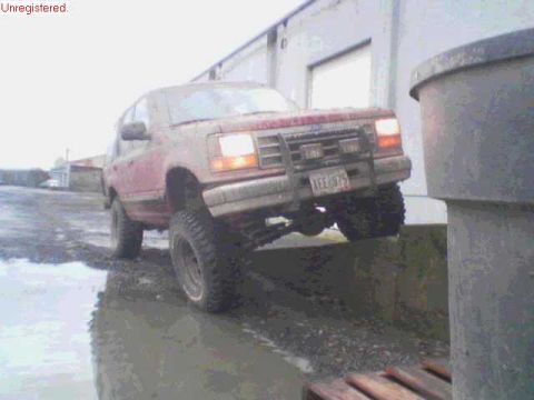 1991 Ford Explorer - Mud-covered red two-door SUV lifted on oversized mud-terrain tires and black steel wheels, climbing a curb next to a metal shop building wi…