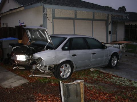 1993 Ford Taurus - A silver 1993 Ford Taurus sedan is parked in a driveway with its trunk open and rear bumper heavily damaged.