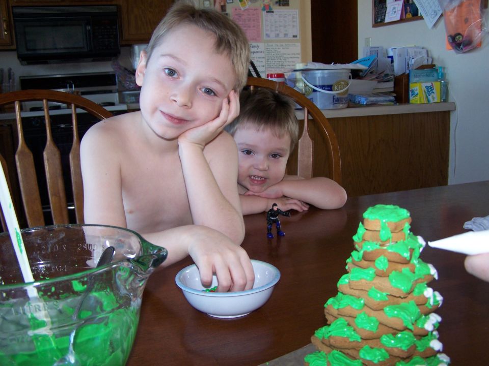 Boy with hand on cheek, another boy behind him, decorating a gingerbread tree with green icing