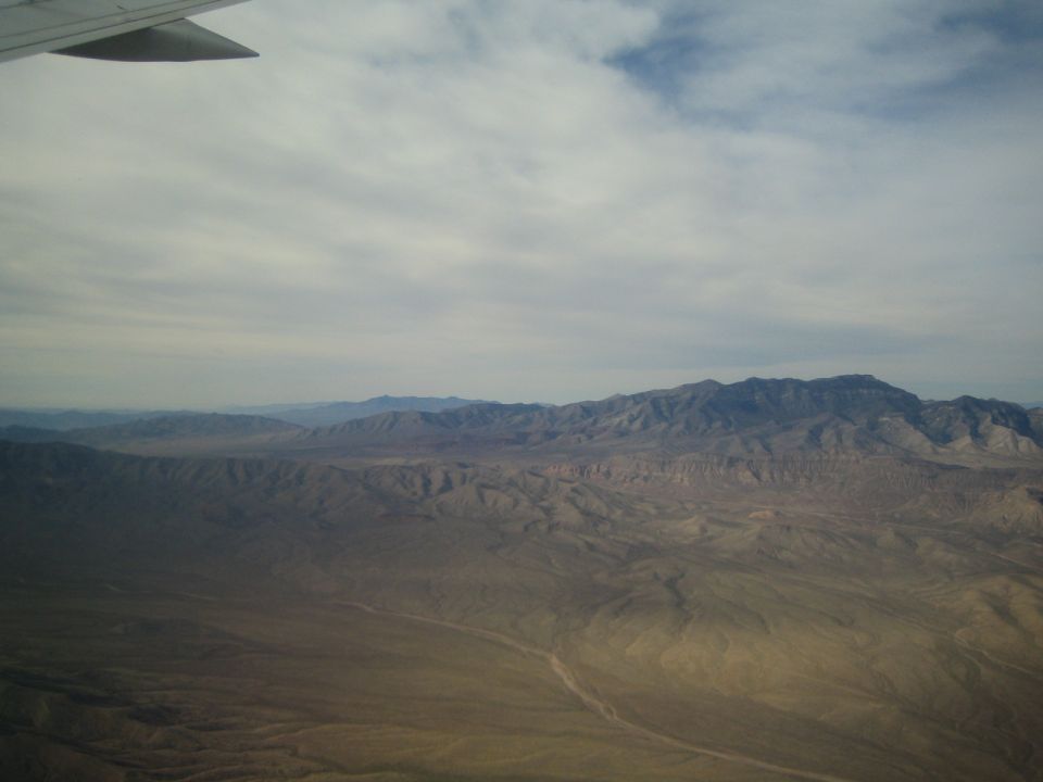 View of a vast desert mountain range from an airplane window, with clouds in the sky