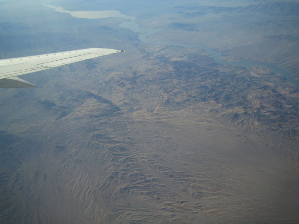 Airplane wing extends over a vast, arid desert landscape with a winding river visible in the distance