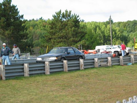1990 Ford Mustang - A dark coupe is on a drag strip with spectators and other vehicles present.
