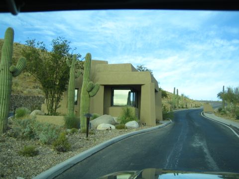 1968 Ford Mustang - A view from a vehicle driving on a desert road towards a modern building, flanked by saguaro cacti under a cloudy sky.