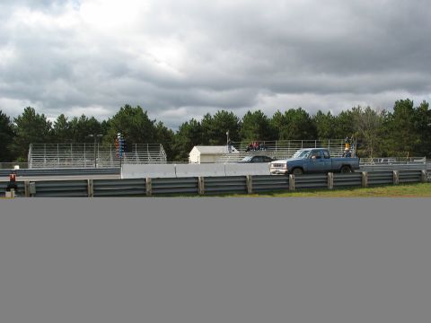 1990 Ford Mustang - Two vehicles are on a race track under a cloudy sky, with trees and spectator stands visible.