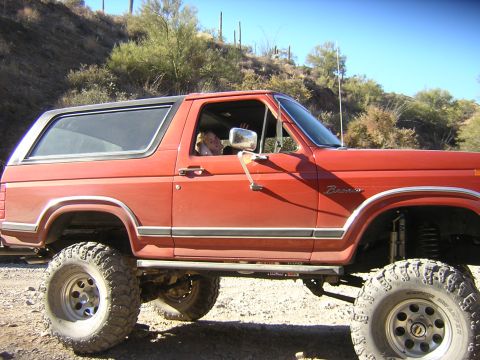 1984 Ford Bronco - Red Ford Bronco with lifted suspension and large tires parked on a dirt road.