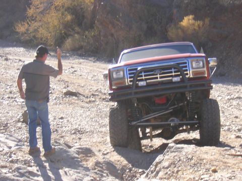 1984 Ford Bronco - A man stands by a modified Ford Bronco as it navigates a rough, rocky terrain.
