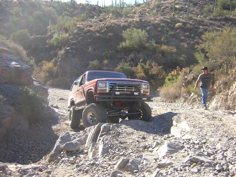 1984 Ford Bronco - A lifted Ford Bronco navigates a rocky, desert-like terrain with a person walking behind it.
