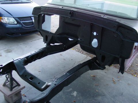 1979 Ford Bronco - Frame and firewall assembly of a vehicle undergoing restoration, with a blue minivan parked nearby.