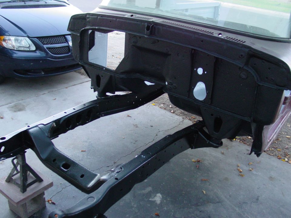 Vehicle frame and firewall assembly on jack stands. Blue minivan in background