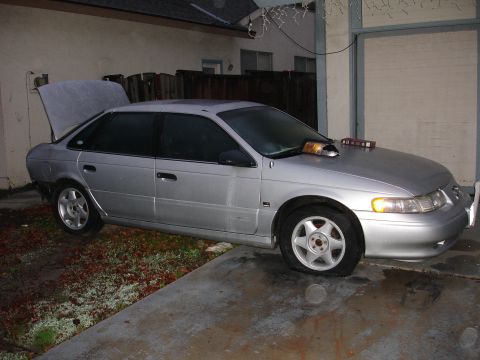 1992 Ford Taurus - Silver 1992 Ford Taurus sedan with a hood scoop and emergency lights, flat front tire.