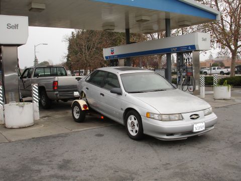 1992 Ford Taurus - A silver 1992 Ford Taurus is being towed by a pickup truck at a gas station, with a trailer attached to the sedan.