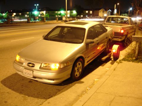 1992 Ford Taurus - A silver 1992 Ford Taurus sedan is being towed by a pickup truck on a city street at night.