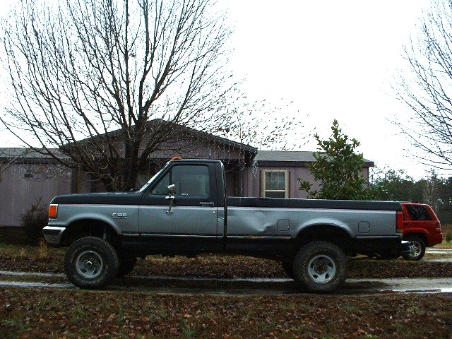 Side view of a lifted 1987 Ford F-150, two-tone gray and black, with body damage