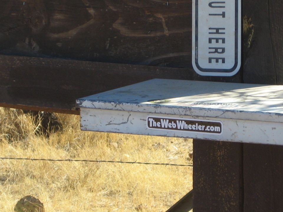 Metal toolbox with 'TheWebWheeler.com' sticker on a wooden post, dry grass and barbed wire in background