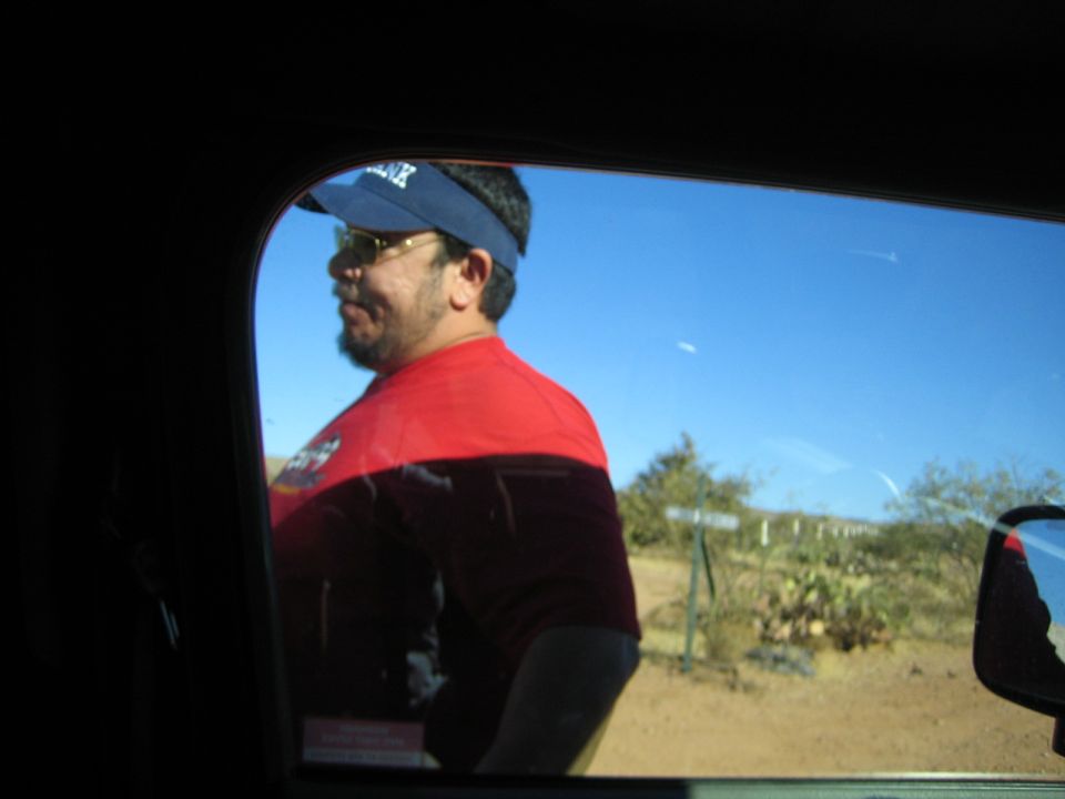 Man in red shirt and blue visor, seen through car window, with desert landscape behind