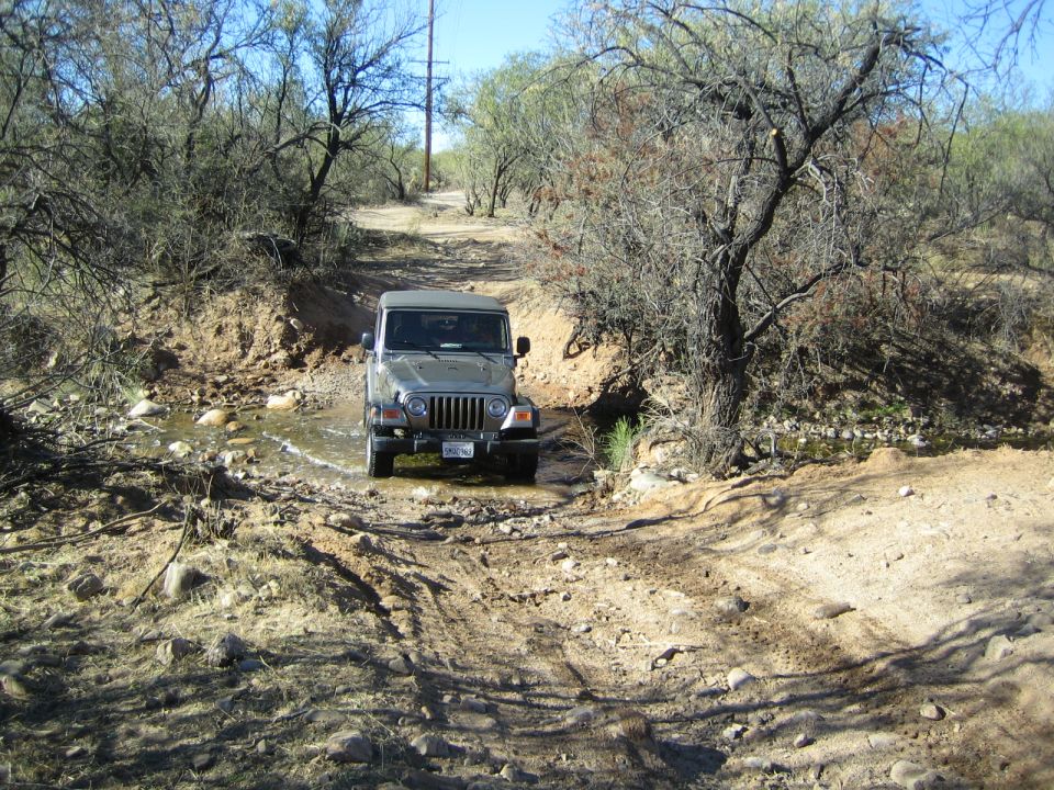 Silver Jeep Wrangler driving on a dirt road with trees and a dry creek bed