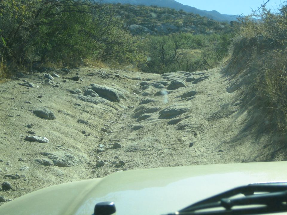View from inside a vehicle driving on a rocky, dirt trail with scrub brush and hills in the background