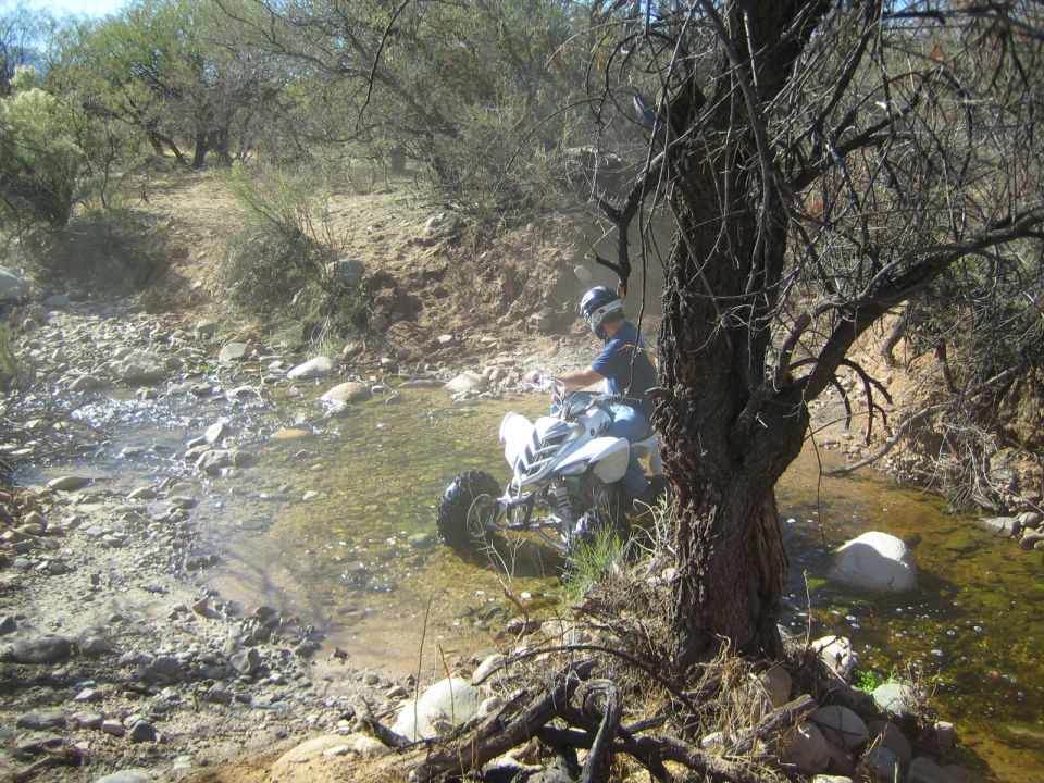 Person on a white ATV rides through a rocky stream bed with trees and brush on the banks