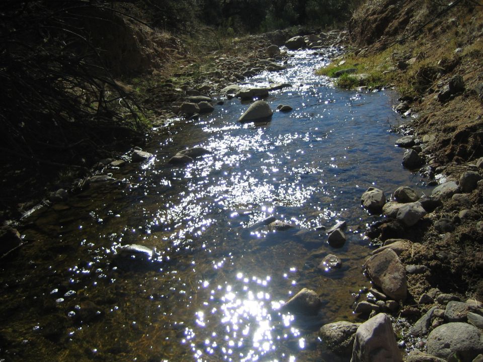Sunlight glints off a shallow stream flowing over rocks in a dry, hilly landscape