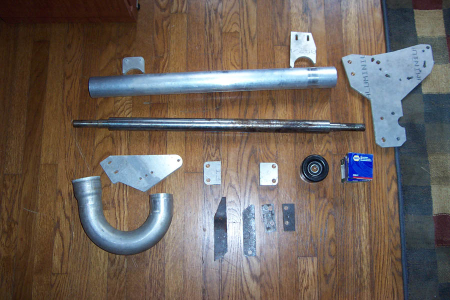 Various metal automotive parts including a large pipe, a shaft, brackets, a pulley, and a blue box on a wooden floor