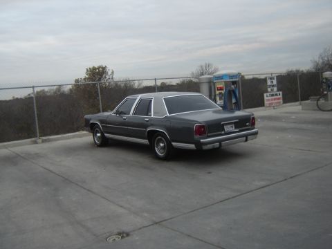 1988 Ford Crown Victoria LTD - A dark sedan is parked at a gas station under an overcast sky.