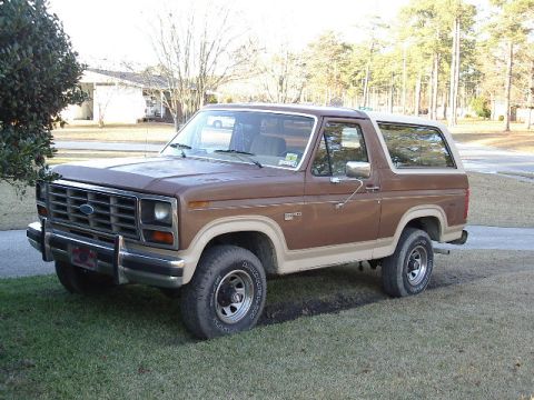 1985 Ford Bronco - This brown two-door Ford Bronco is parked on a grassy area with trees in the background.