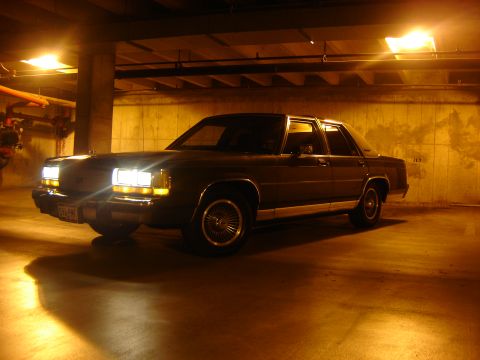 1988 Ford Crown Victoria LTD - A dark sedan is captured in a garage at night, its headlights illuminating the concrete floor.