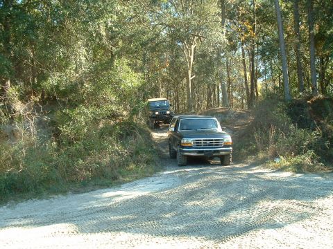 1995 Ford Bronco - A black 1995 Ford Bronco navigates a dirt path through a wooded area, followed by a Jeep.