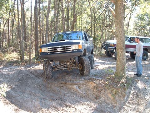 1995 Ford Bronco - A lifted Ford F-Series truck with large off-road tires is parked in a dirt lot with another vehicle and a man nearby.