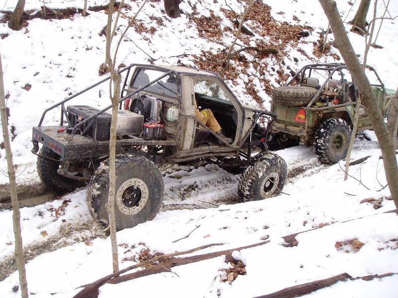 Two heavily modified off-road vehicles with large tires navigate a snowy, wooded trail