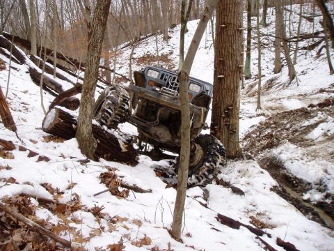1992 Ford Bronco - An off-road vehicle navigates a challenging snowy terrain, tilted precariously on logs and mud.