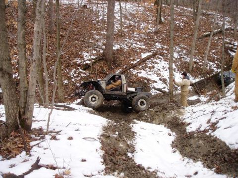1992 Ford Bronco - A heavily modified off-road vehicle navigates a challenging muddy and snowy trail in a wooded environment.