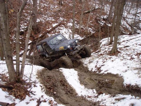 1992 Ford Bronco - A dark Jeep Wrangler navigates a challenging muddy and snowy terrain on a wooded slope.